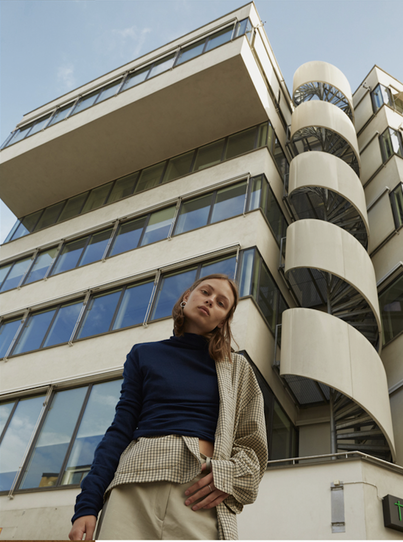image of a swedish female model wearing a blue sweater, beige checked shirt, and beige pants standing in front of a parking garage