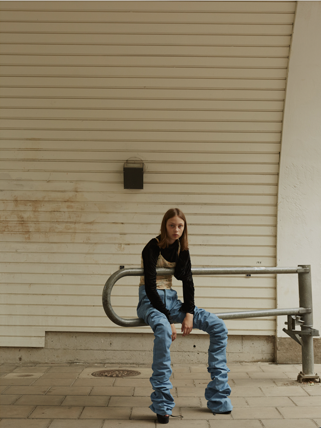 image of a swedish female model wearing extra long jeans scrunched up, a black sweater, and a printed cami sitting on an industrial gate