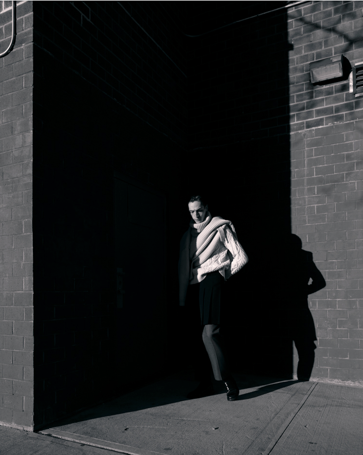 image of a male model in black and white wearing two cable knit sweaters, trousers, and loafers in between two buildings outside