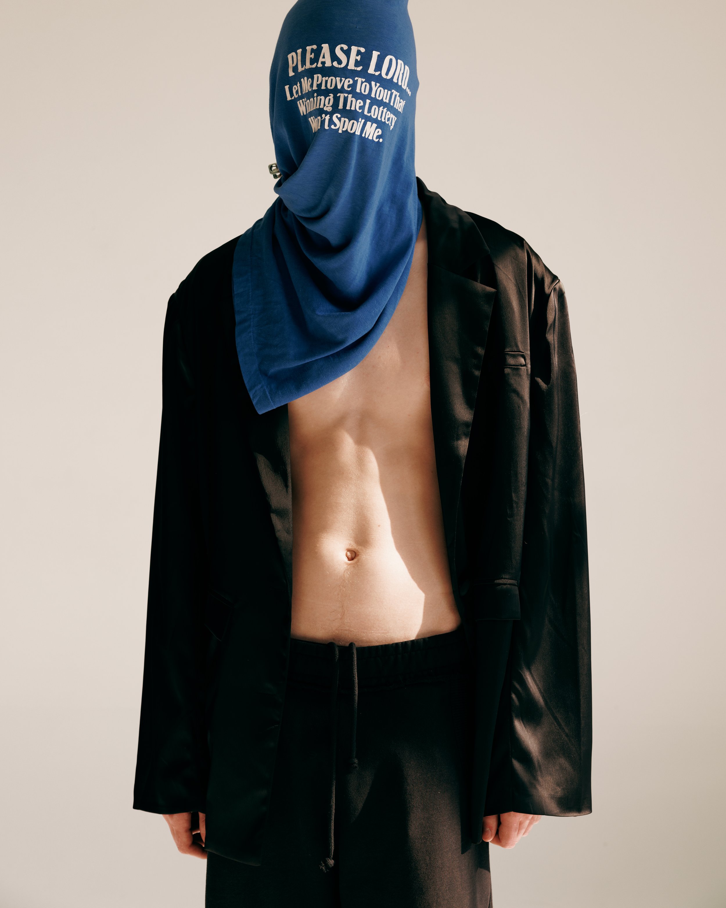 portrait of a young redheaded white male model with a shirt over his head that reads "please lord let me prove to you that winning the lottery won't spoil me" whith a shiny black blazer and black sweatpants in a white studio