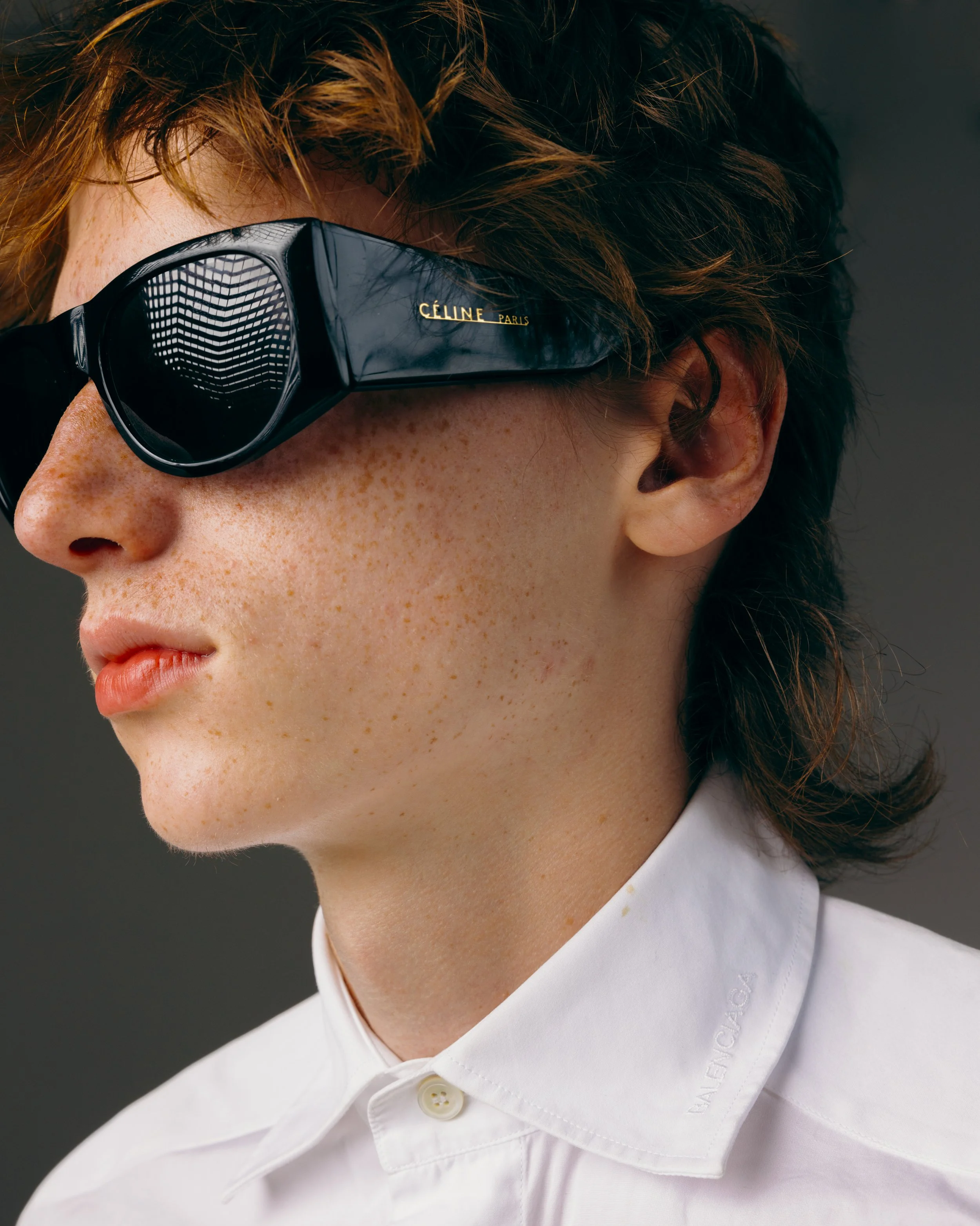 portrait of a redheaded white male model with freckles wearing large black Celine sunglasses and a white balenciaga shirt in a white studio