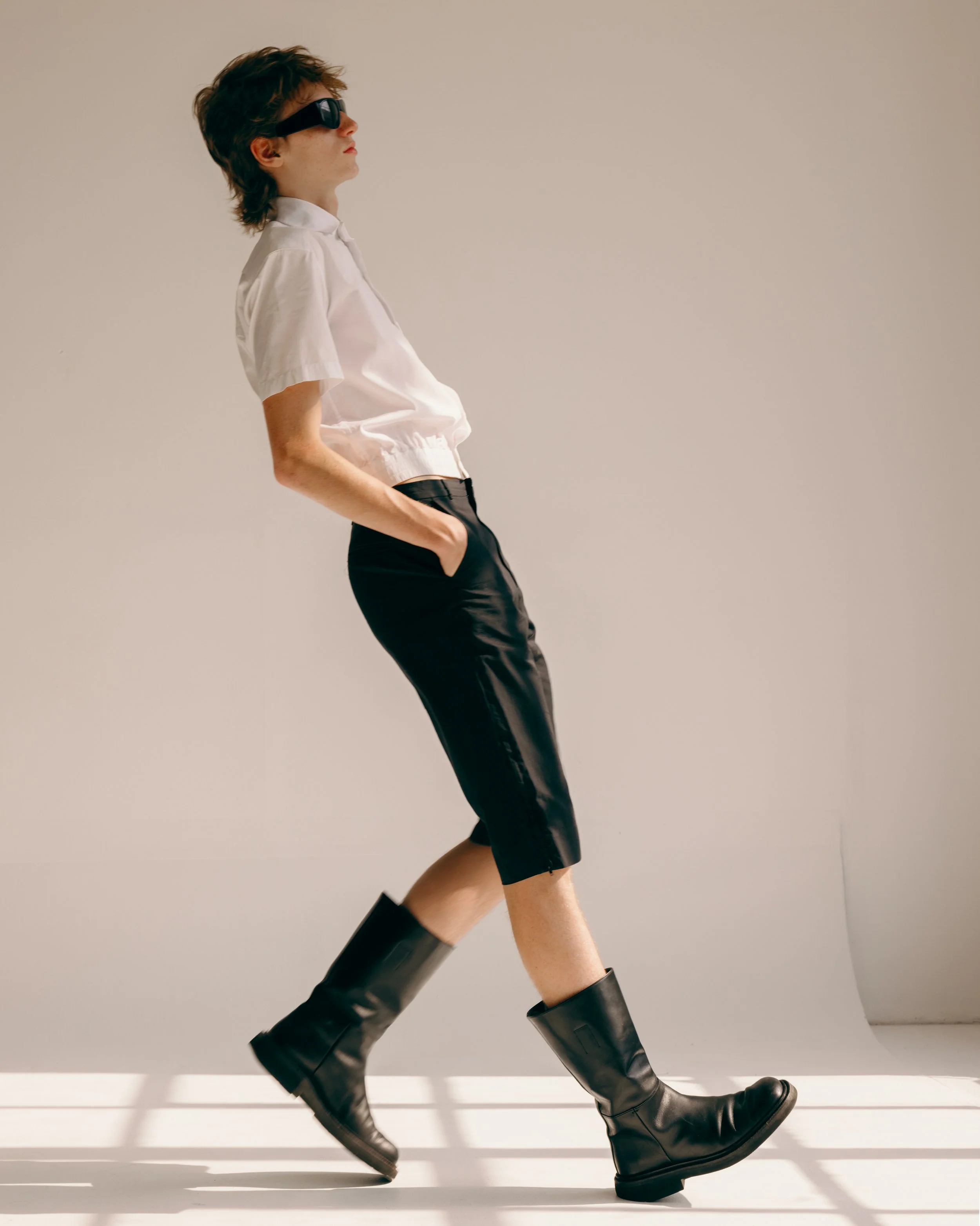 image of a readheaded white male model wearing a white short sleeve balenciaga shirt, black knee length shorts, and tall black boots falling backwards in a white studio