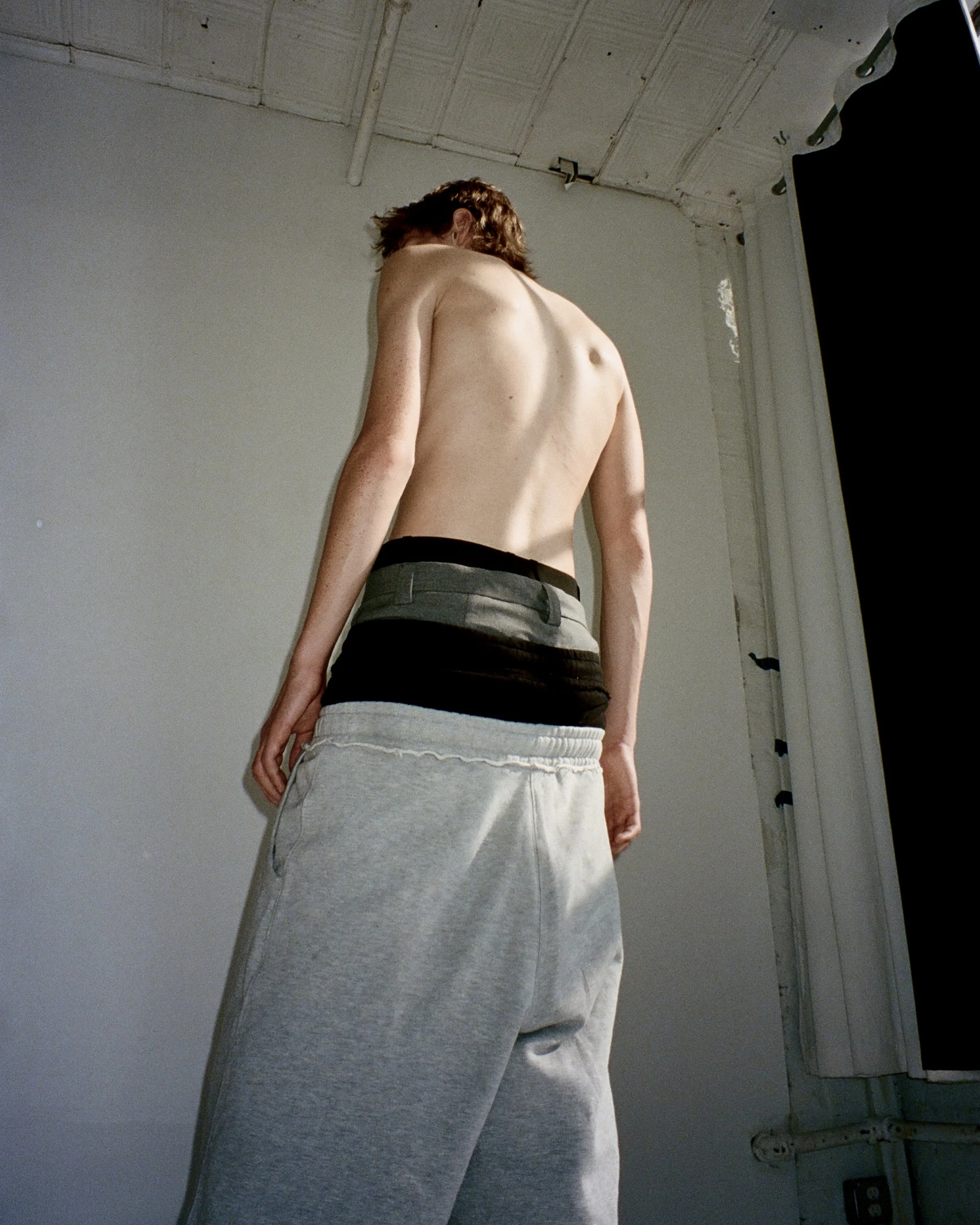 portrait of a white male model from behind wearing 4 pairs of pants stacked on top of each other in a white studio
