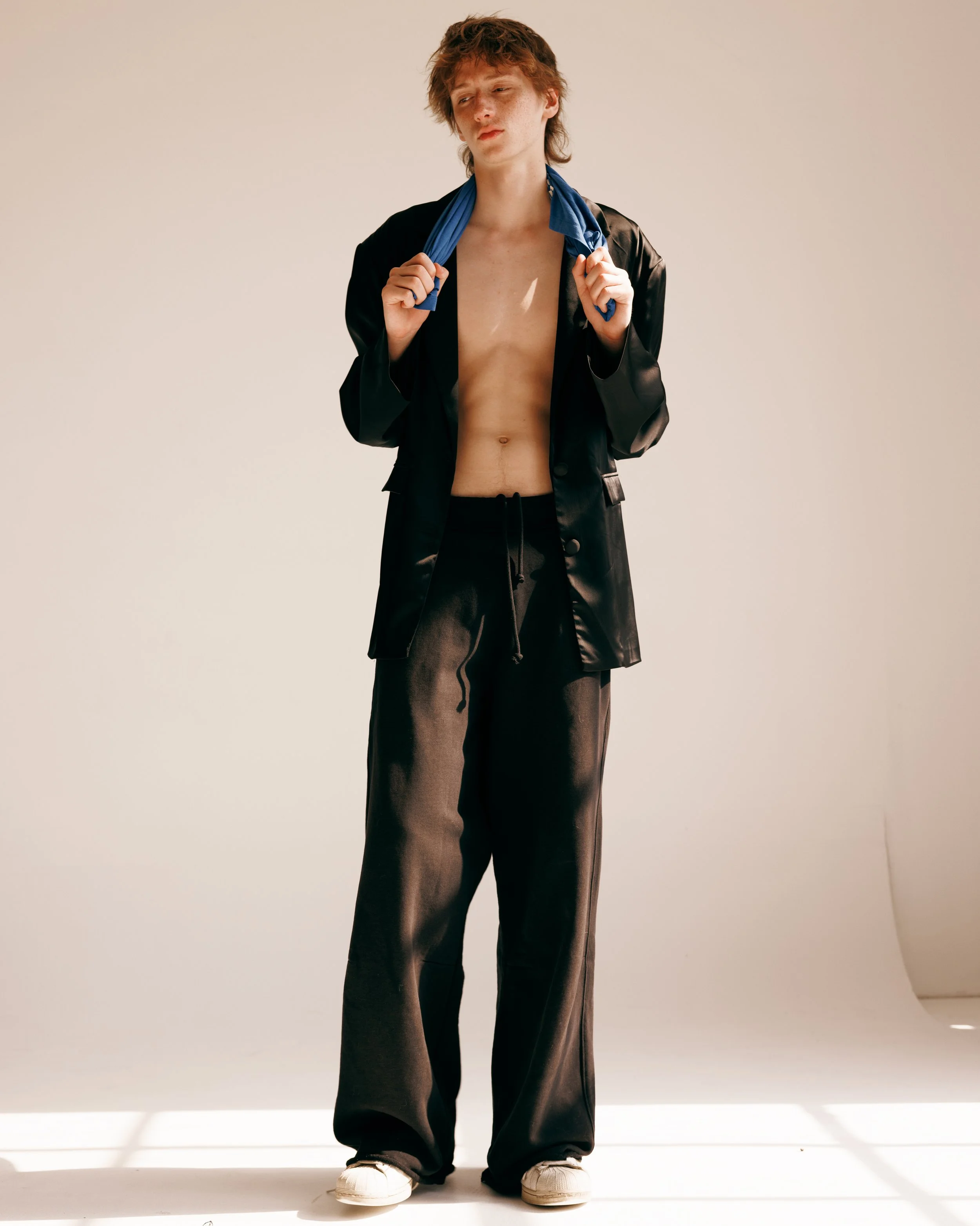 portrait of a redheaded white male model wearing a shiny black blazer, black sweatpants, and dirty white sneakers, holding a blue shirt behind his neck