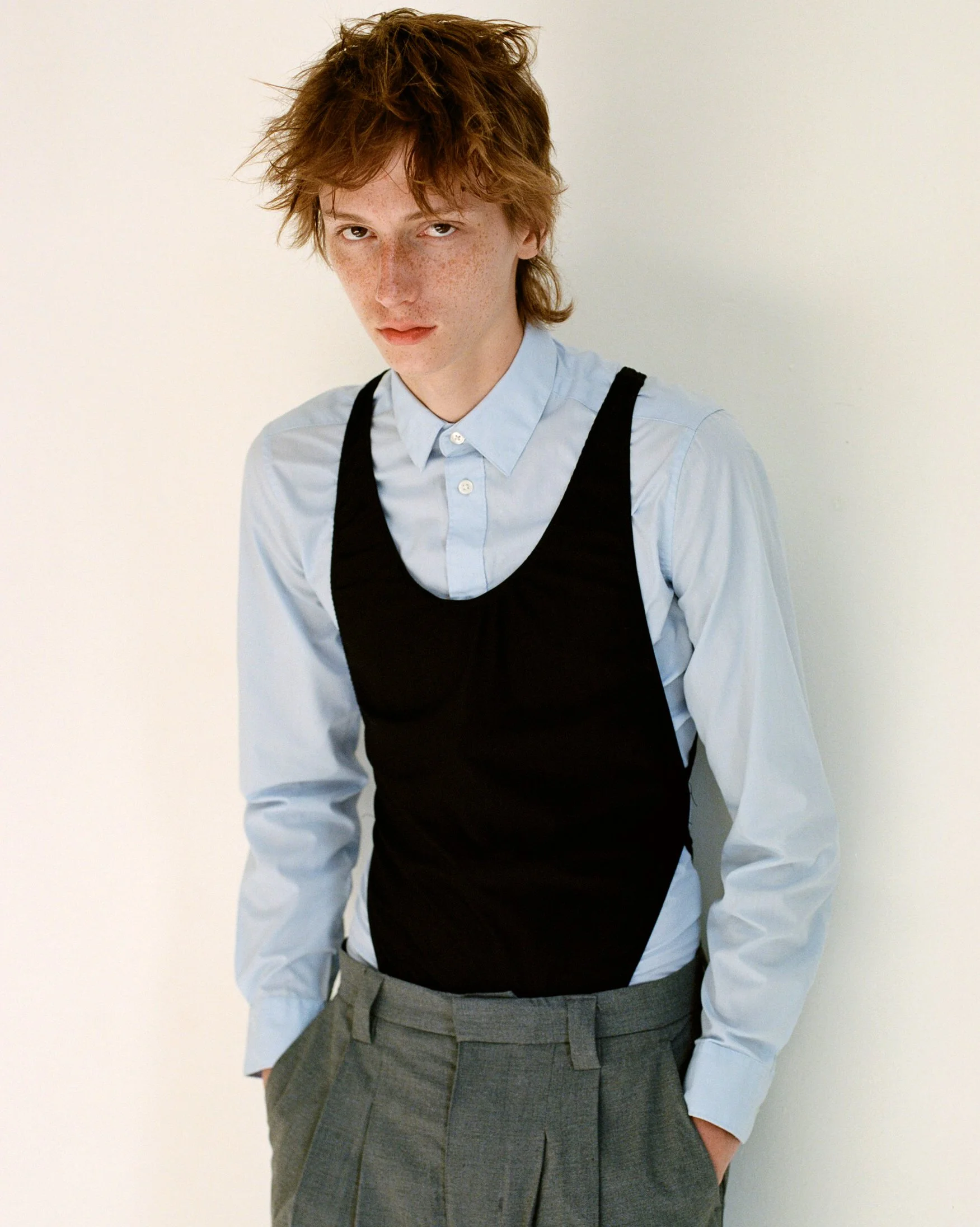 portrait of a redheaded white male model wearing a blue button down shirt with a black leotard on top and grey trousers on a white background