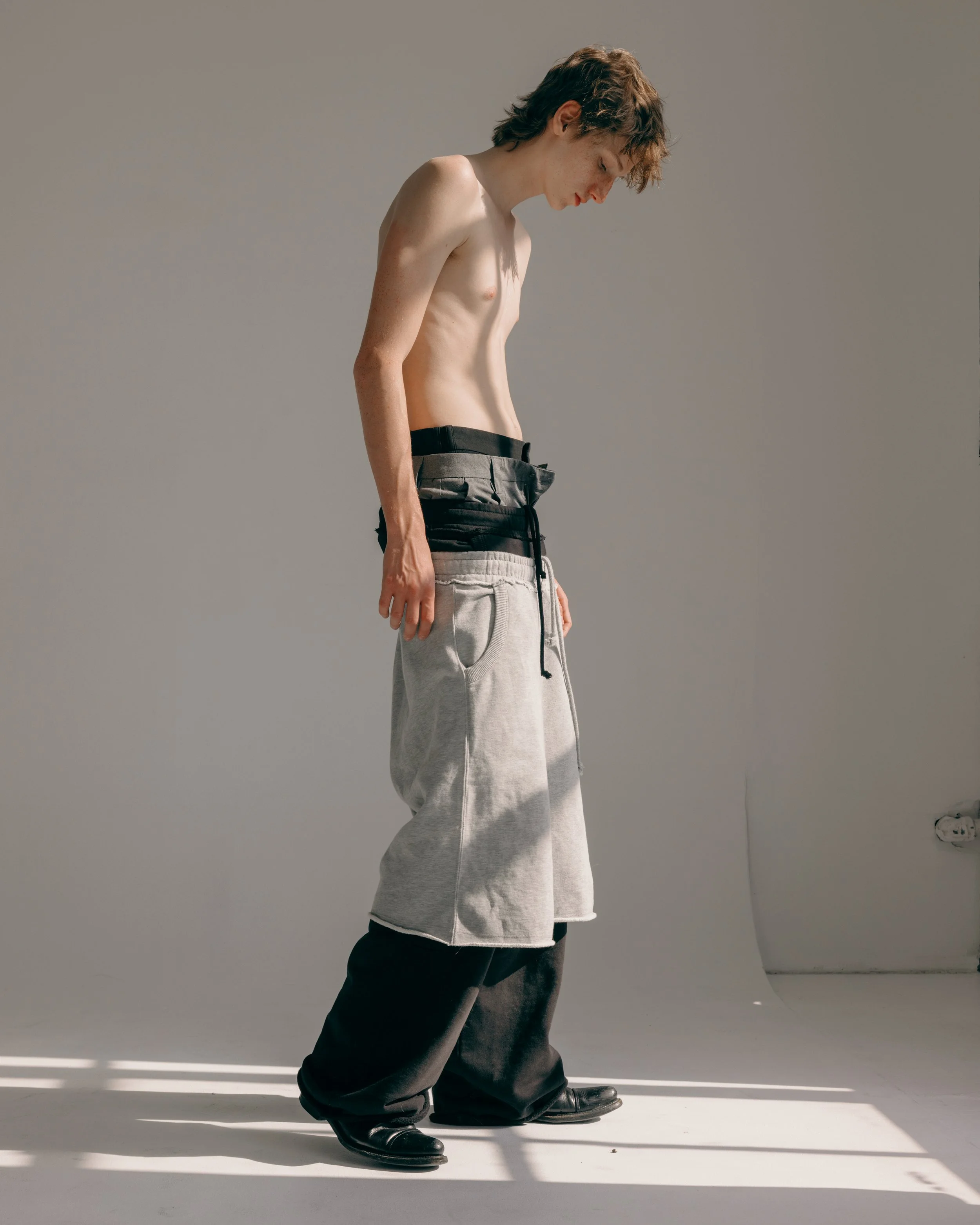 image of a redheaded white male model wearing 4 pairs of pants stacked on top of one another and black boots in a white studio