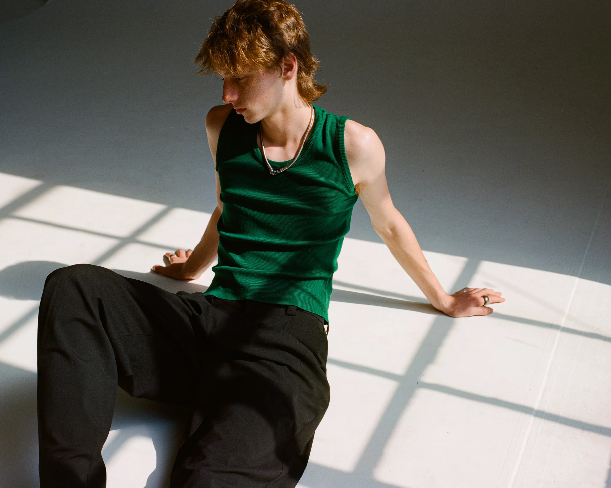 portrait of a redheaded white male model sitting on the floor of a white photo studio in the sunlight wearing a green tank top, black pants, and a silver necklace