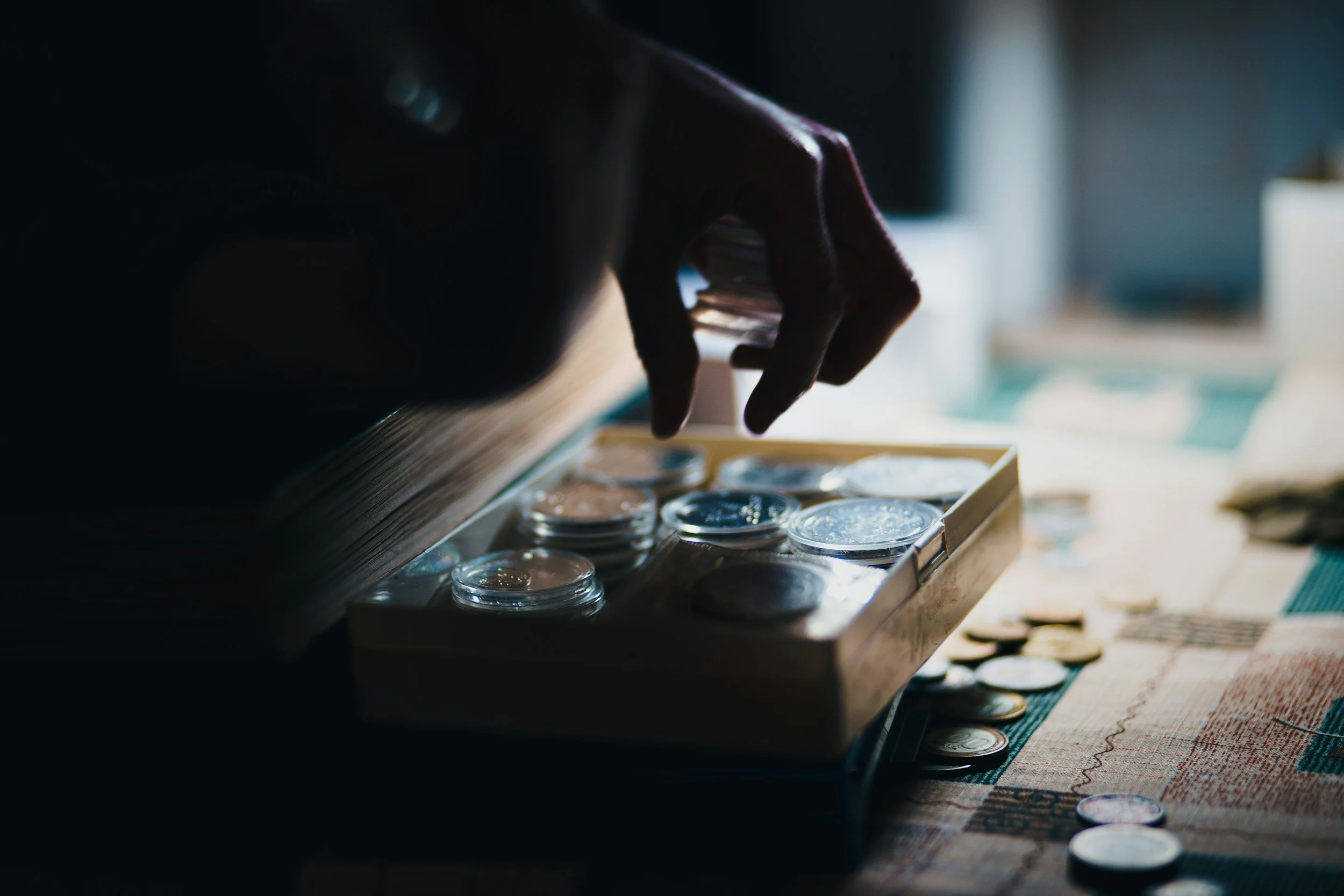A person's hand reaching into a wooden box of assorted coins, with some coins scattered on a table.