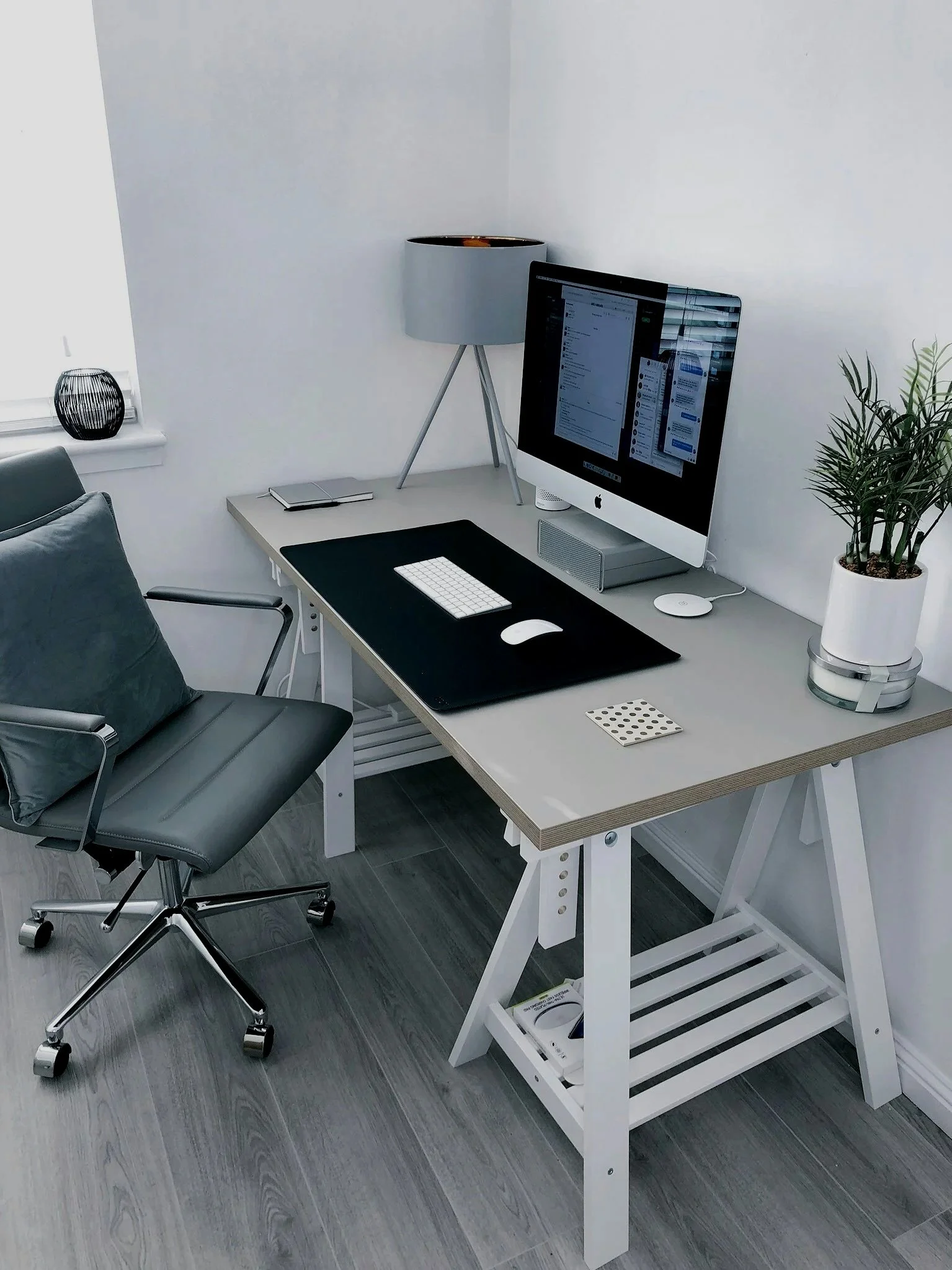 Home office setup with a desk, an iMac computer, a black desk mat, a wireless keyboard and mouse, a desk lamp, a potted plant, and a black decorative vase on the windowsill.