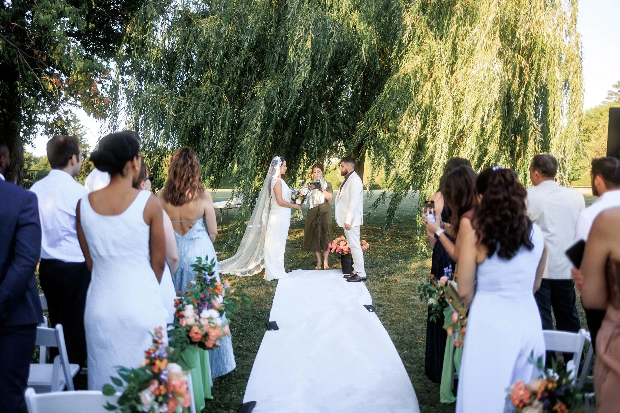 Outdoor wedding ceremony with a couple exchanging vows under a large tree, surrounded by seated wedding guests.