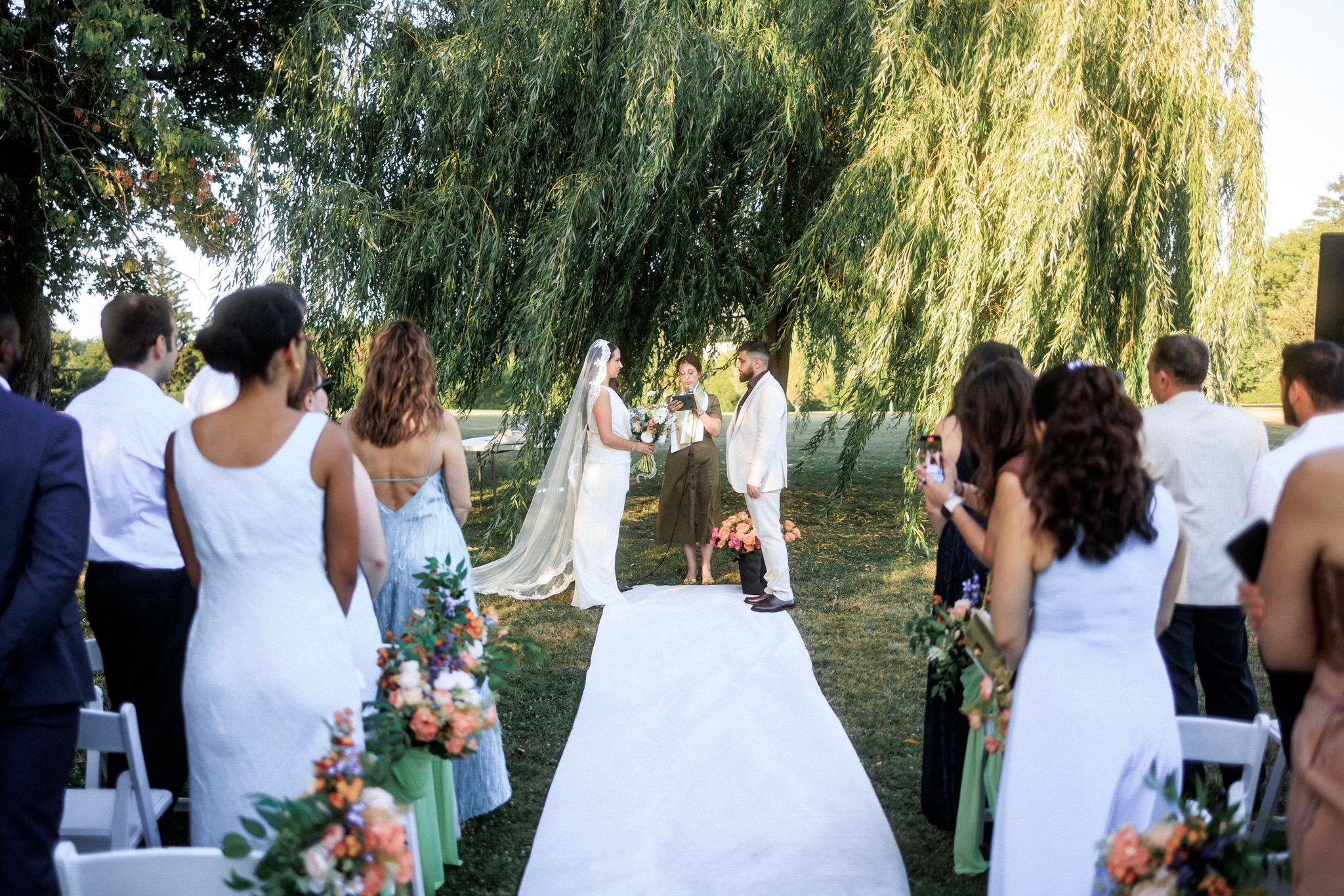 A wedding ceremony taking place outdoors under a large tree, with a bride and groom facing each other on a white aisle, surrounded by guests watching and taking photos. The bride is holding a bouquet and wearing a long veil, and the groom is in a white suit. An officiant is standing in front of them.