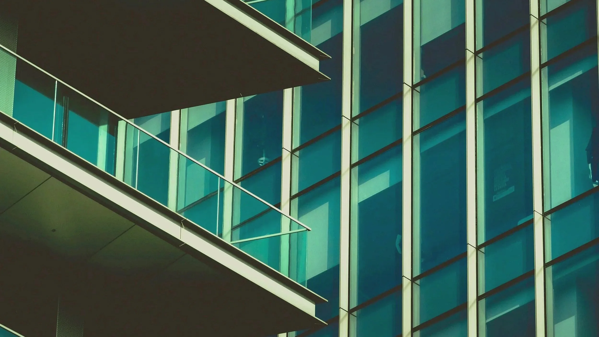 Modern building with glass windows reflecting sunlight, featuring balconies, and a person visible inside through the window.