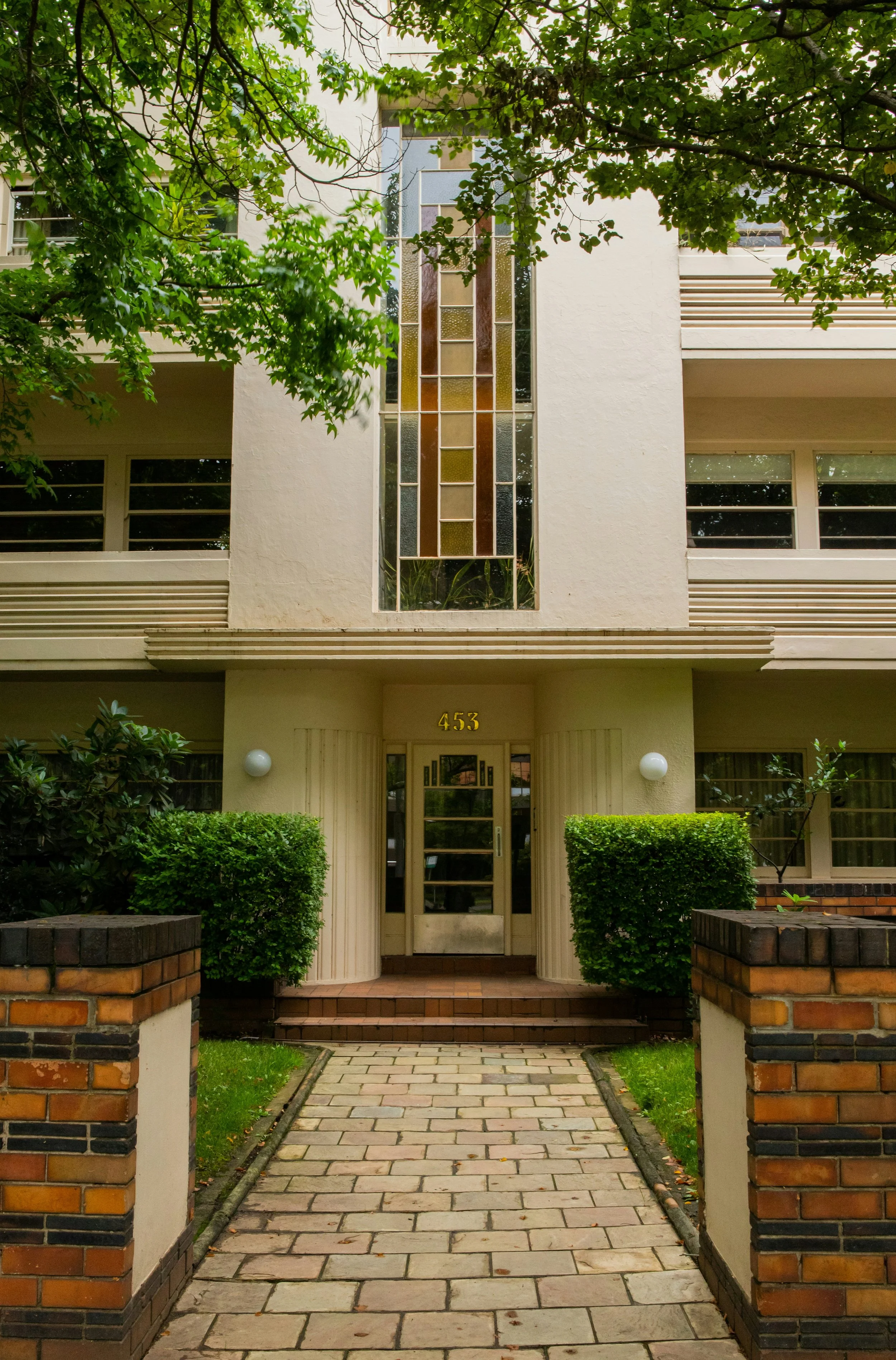 Front entrance of a residential building with the address 453 displayed above the door. The entrance is flanked by green bushes and brick pillars, with a brick walkway leading to the door. There are leafy green trees visible above.