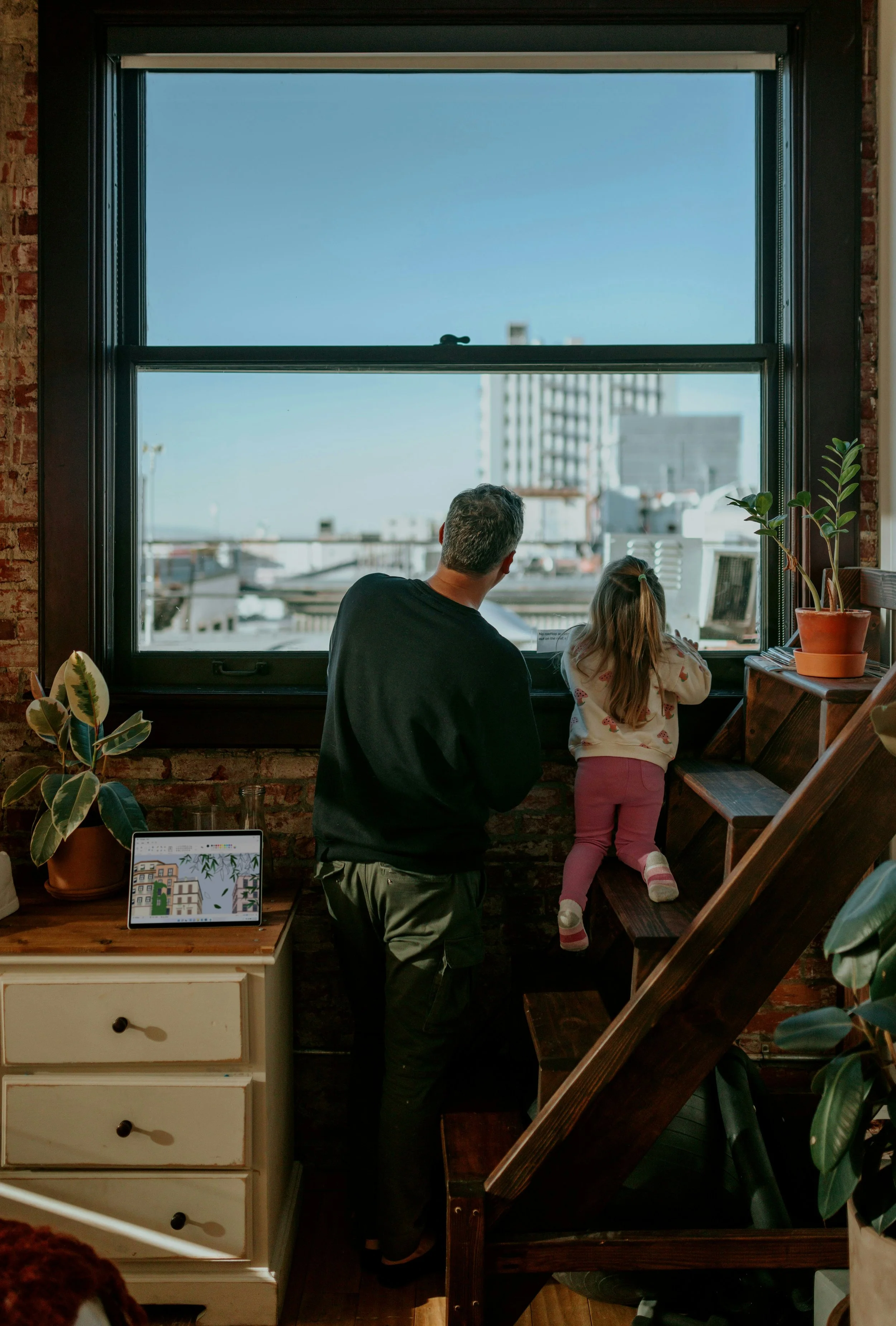 A man and a young girl looking out a large window in an urban apartment, with city buildings outside. The girl is standing on a staircase, and the man is standing beside her. There are potted plants and a tablet on a nearby table inside the apartment.