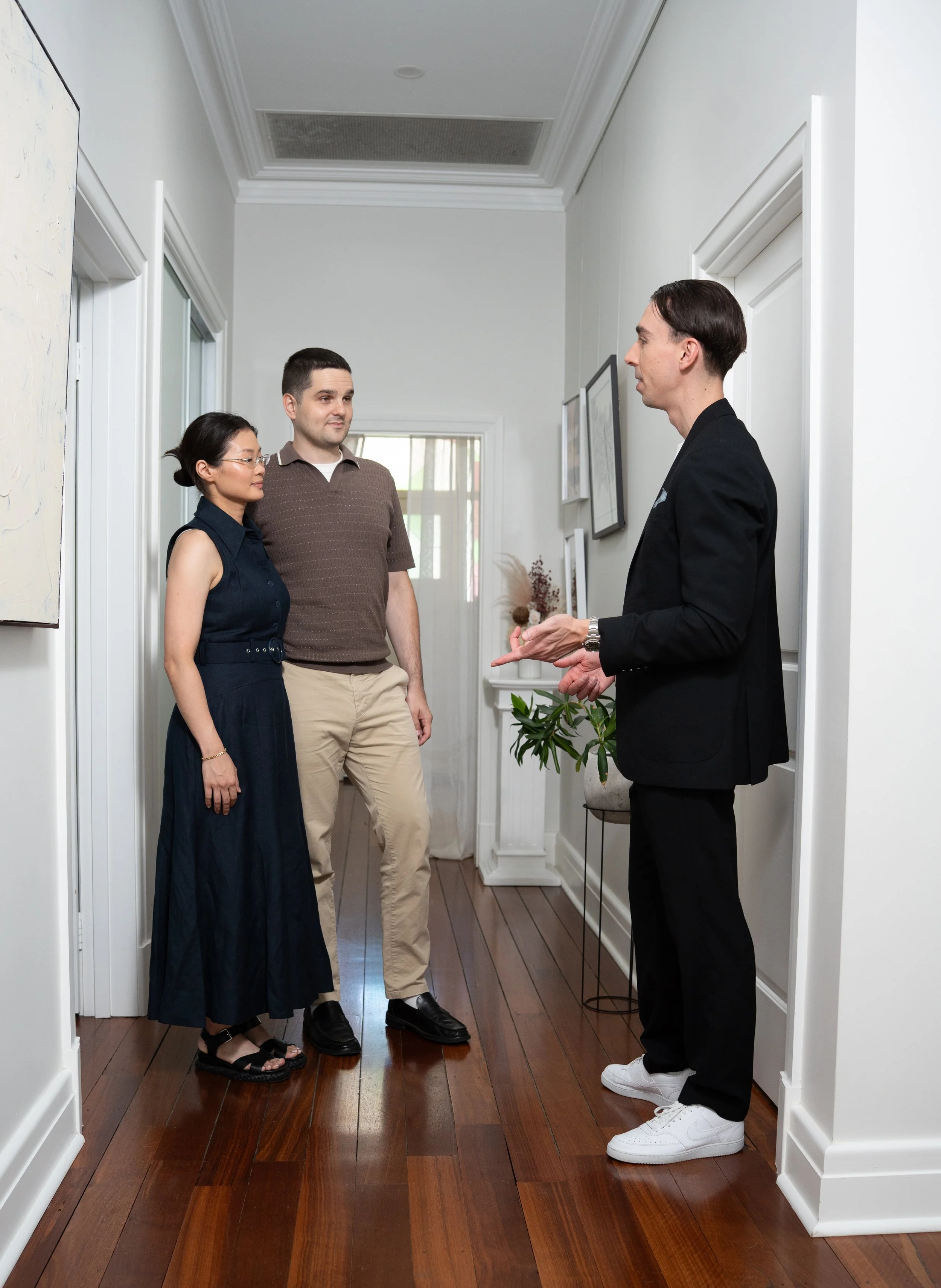 A real estate agent in a black suit and white sneakers is speaking to a couple inside a home, with the couple listening attentively. The interior has white walls, wooden floor, and framed pictures on the wall.