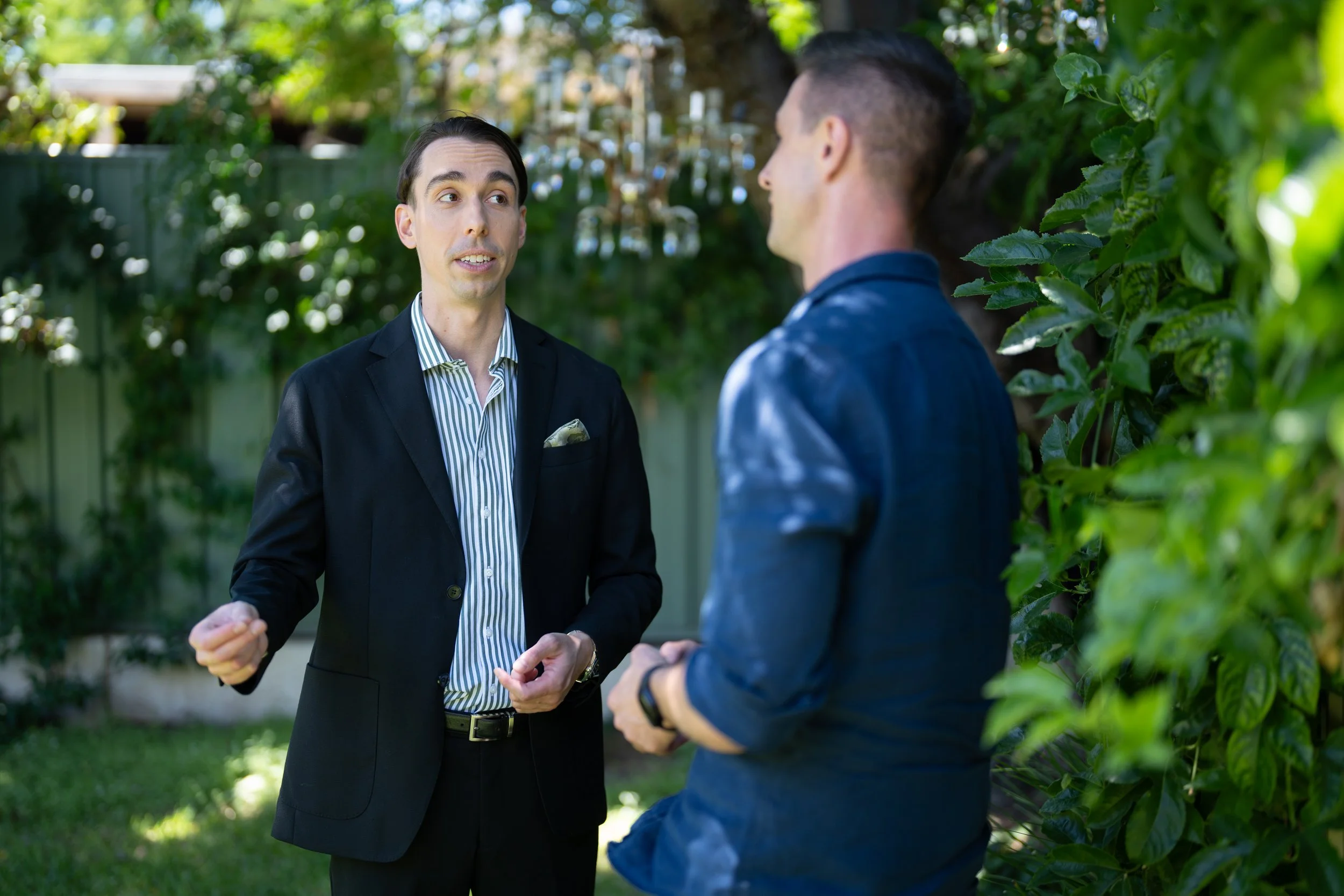 Two men having a conversation outdoors near a green fence with trees and plants in the background.
