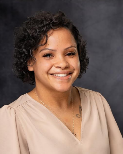 A woman with dark hair smiling outdoors near a tree, wearing a white shirt.