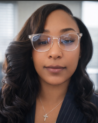 A woman with long black wavy hair wearing a blue collared shirt and hoop earrings, smiling with a neutral dark background.