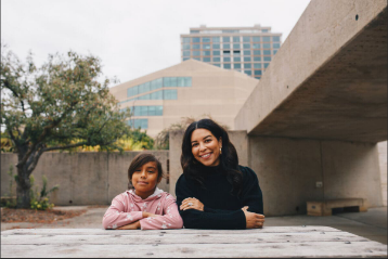 A woman and a young girl sitting on a concrete surface outdoors, with urban buildings and a tree in the background.