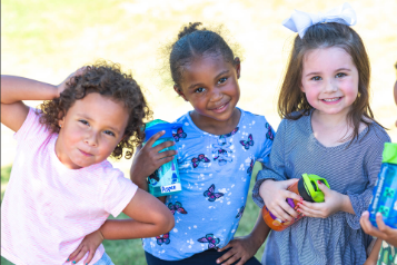 Three young girls smiling outdoors, one holding a toy cellphone and another holding a toy in a grassy area.