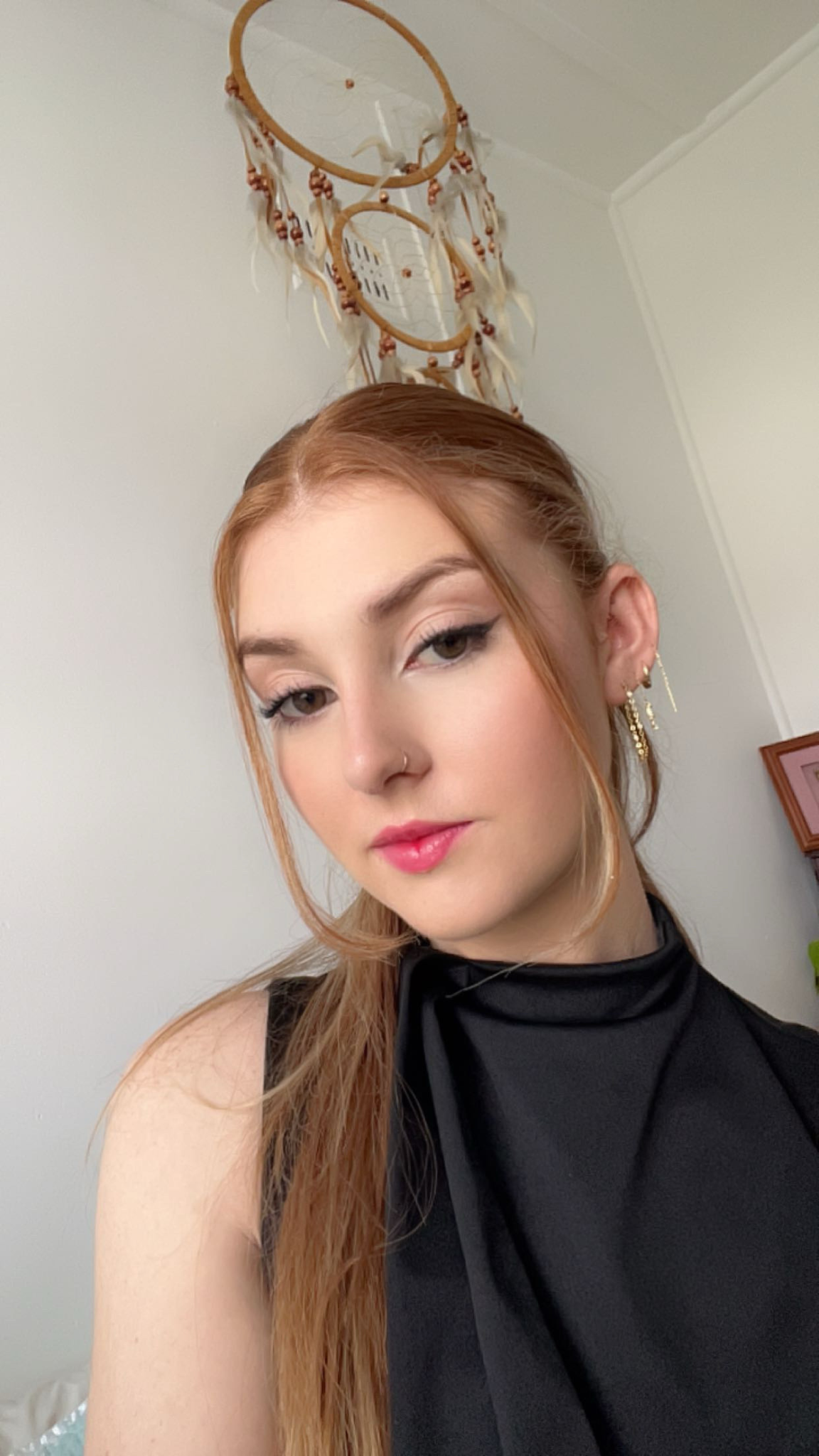 A young woman with red hair and earrings taking a selfie in a room with a dreamcatcher hanging on the wall behind her.