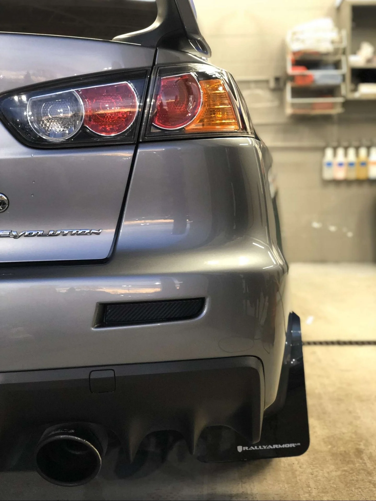 Close-up of the rear left side of a silver Mitsubishi Lancer Evolution, showing the tail light, rear bumper, and a black Rally Armor mud flap in a garage with shelves and bottles in the background.