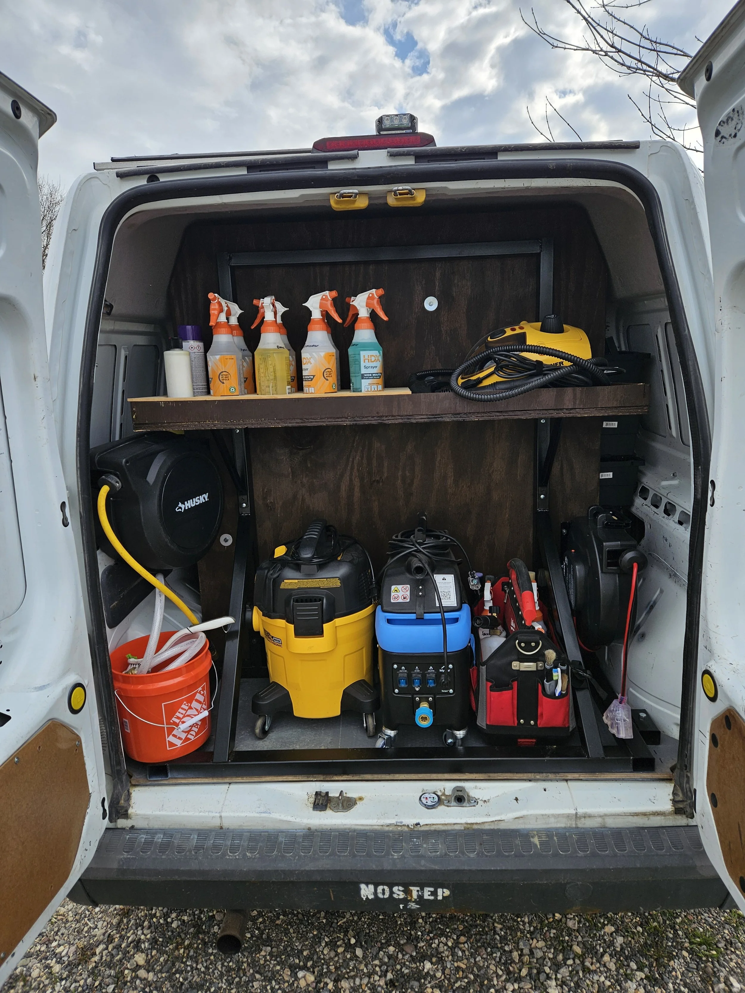 The back of a work van with shelves containing cleaning supplies, a wet/dry vacuum, a toolbox, and other tools organized inside.