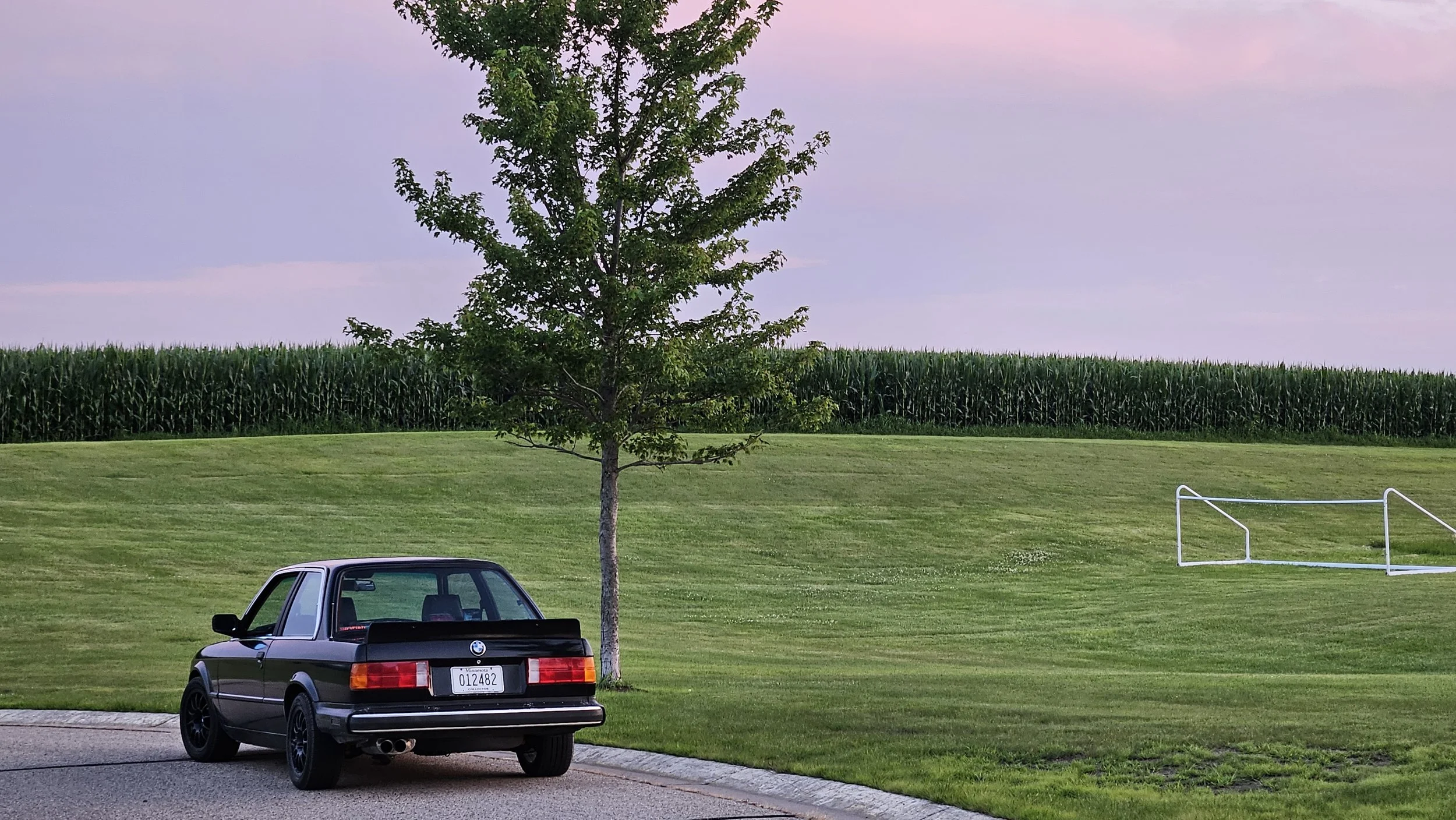A black BMW E30 parked on the side of a grassy area with a tree in front, a field of tall corn in the background, and playground equipment on the right, under a pinkish-purple sky.