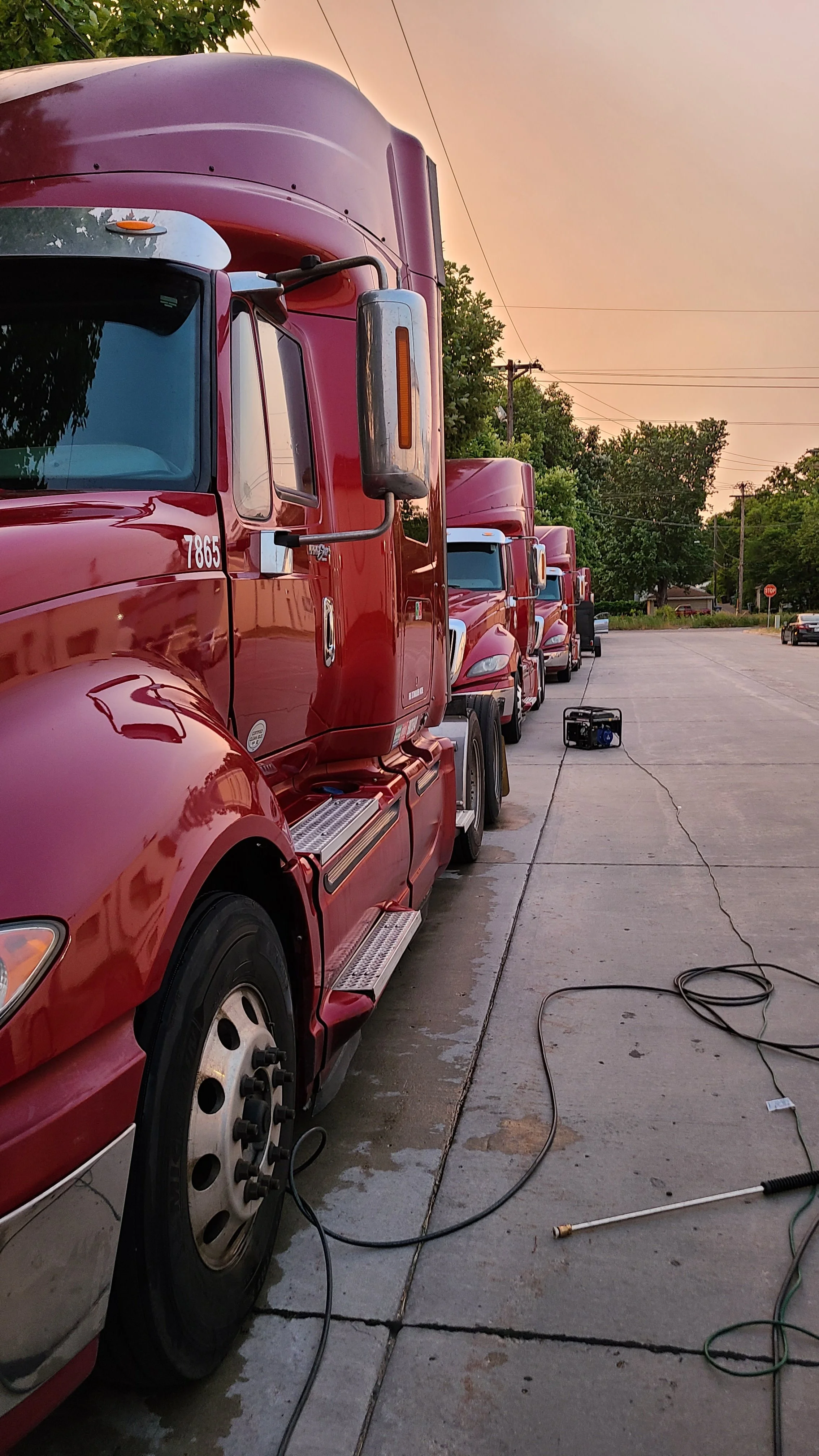 A row of red semi-trucks parked on a sidewalk during sunset with a portable generator and power cords on the ground.
