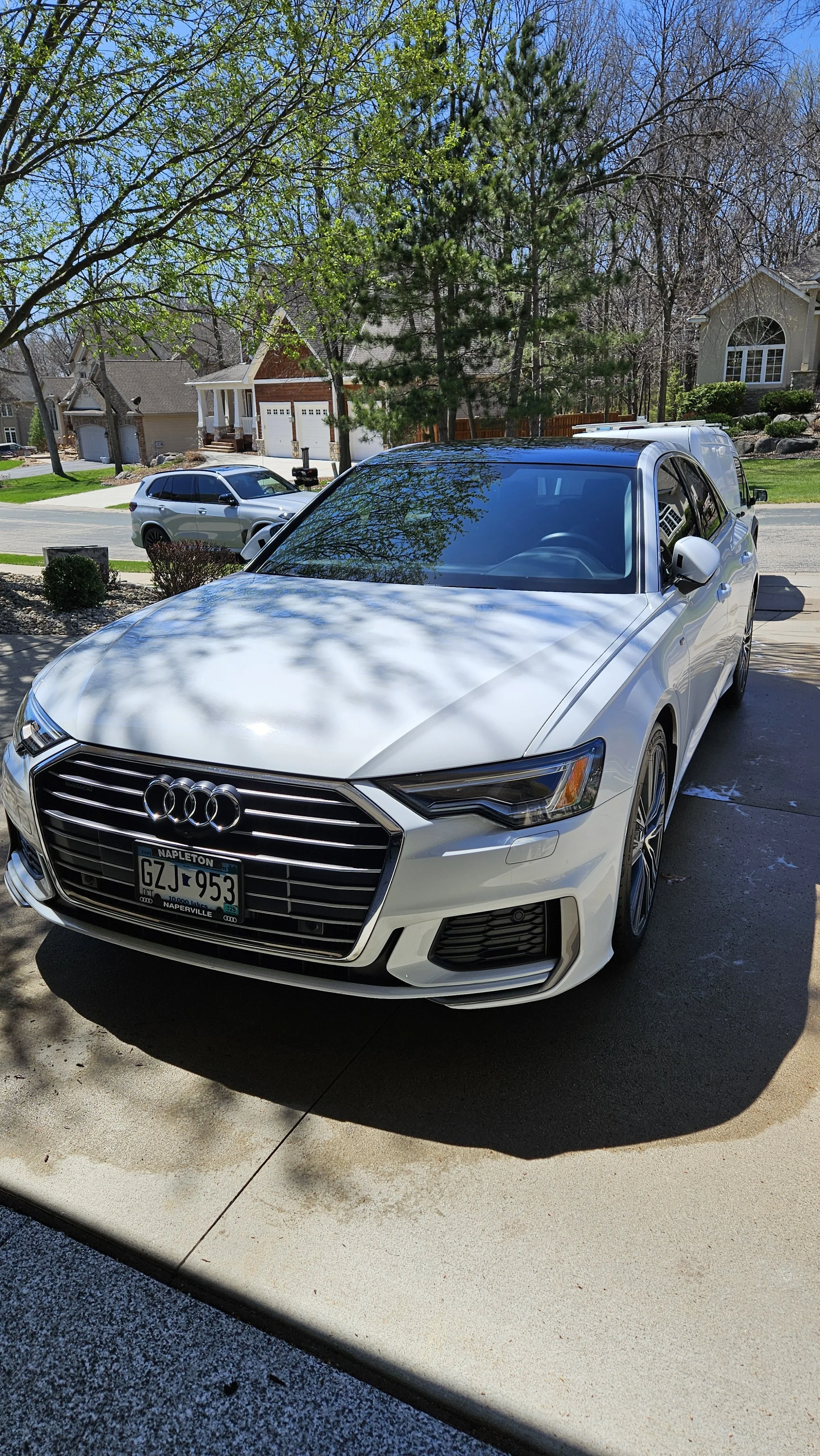 A white Audi sedan parked in a driveway with tree shadows on it, in a suburban neighborhood with houses and trees visible in the background.