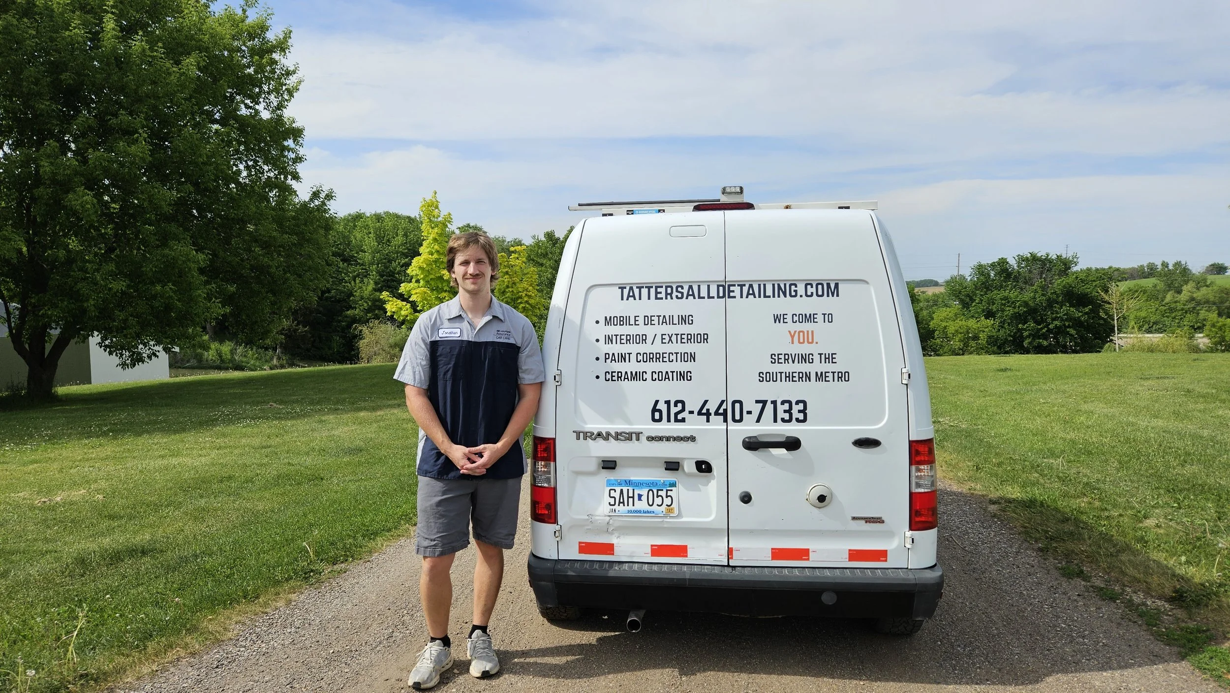 A man standing next to a white van with the website tattersalldetailing.com printed on it, in a grassy area with trees and a blue sky in the background.