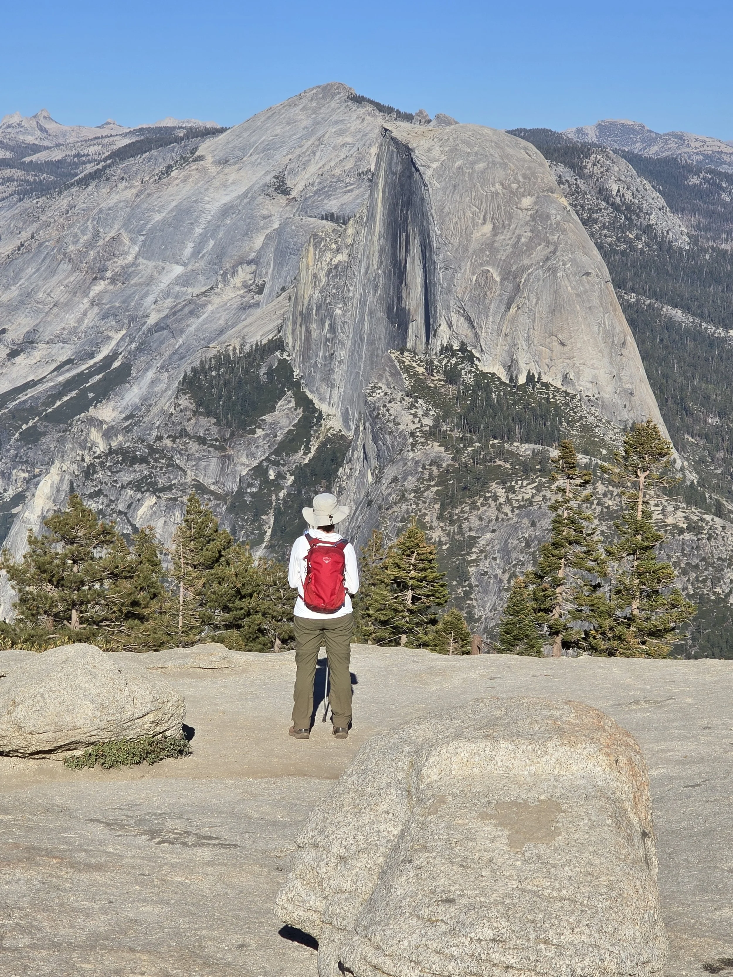 Hiker standing on a rocky trail overlooking a large granite mountain with a sheer cliff face, surrounded by pine trees and distant mountain peaks under a clear blue sky.