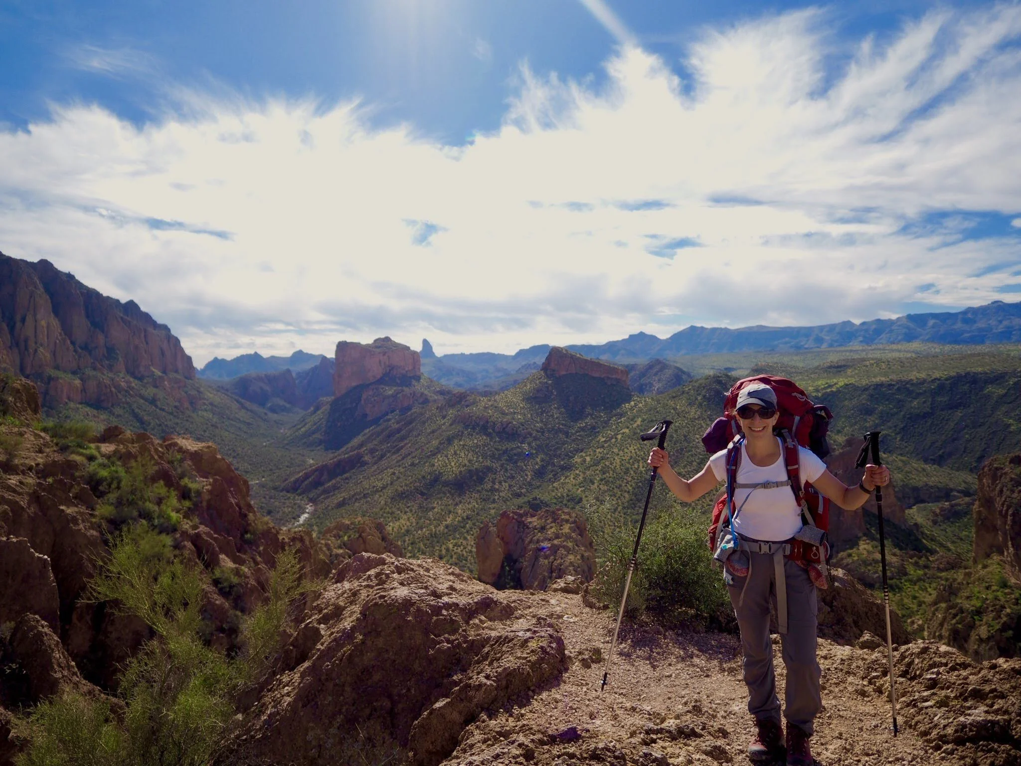 A woman smiling with sunglasses, wearing a white t-shirt and a large backpack, holding trekking poles on a trail in a mountainous landscape with rocky peaks and green vegetation under a partly cloudy sky.