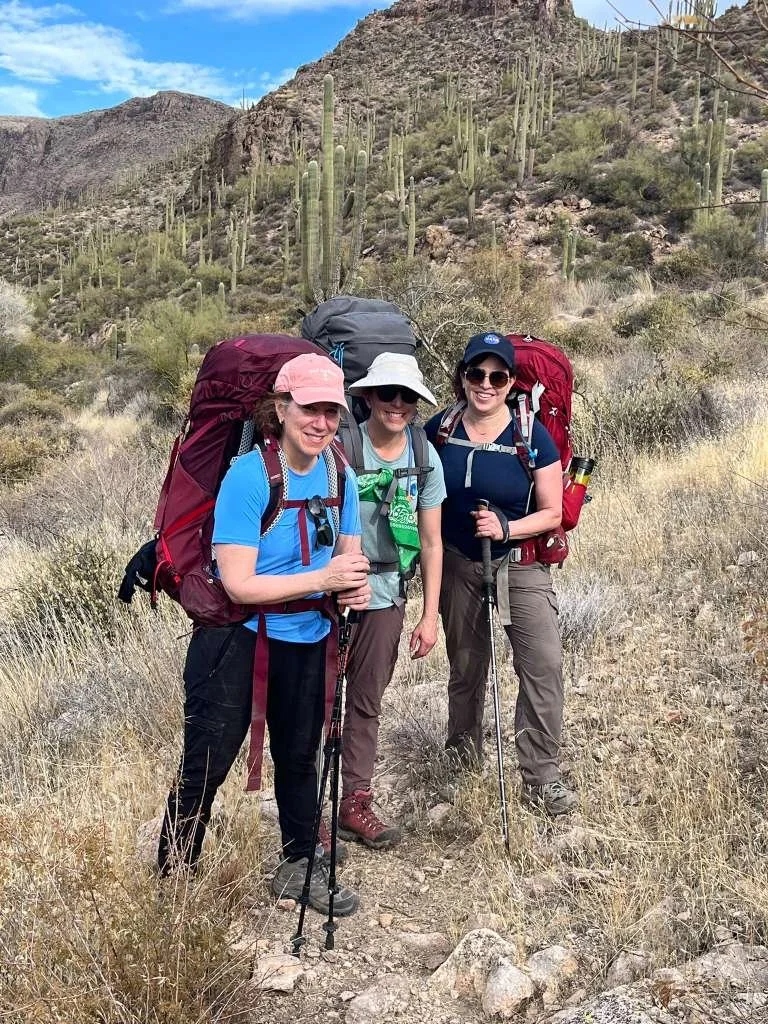 Three women with backpacks and hiking poles on a desert trail surrounded by cacti and dry shrubbery, mountains in the background under a partly cloudy sky.