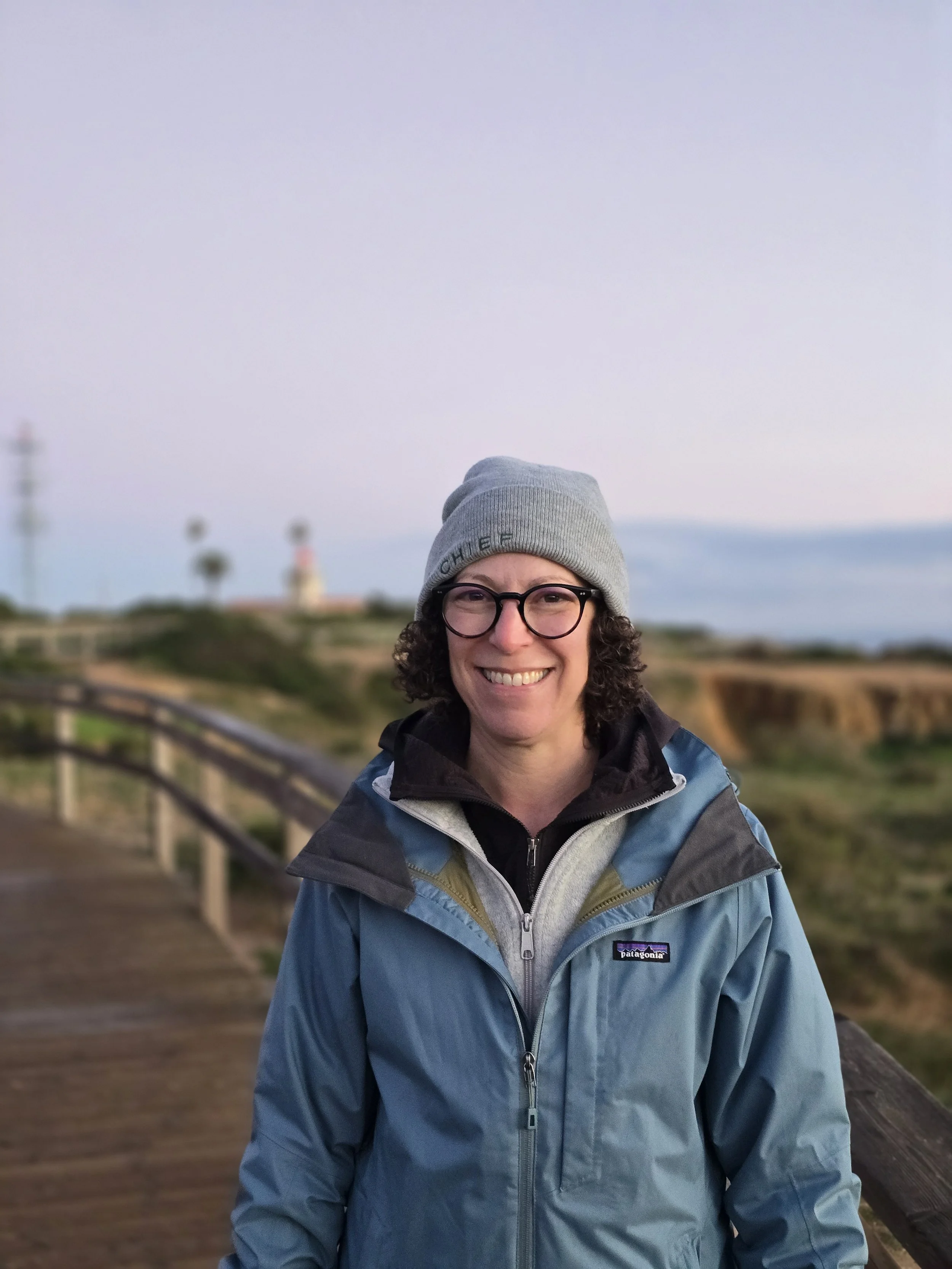 A smiling woman with glasses wearing a gray beanie and a blue Patagonia jacket, standing outdoors near a wooden railing with a blurred background of a coastline, lighthouse, and blue sky.