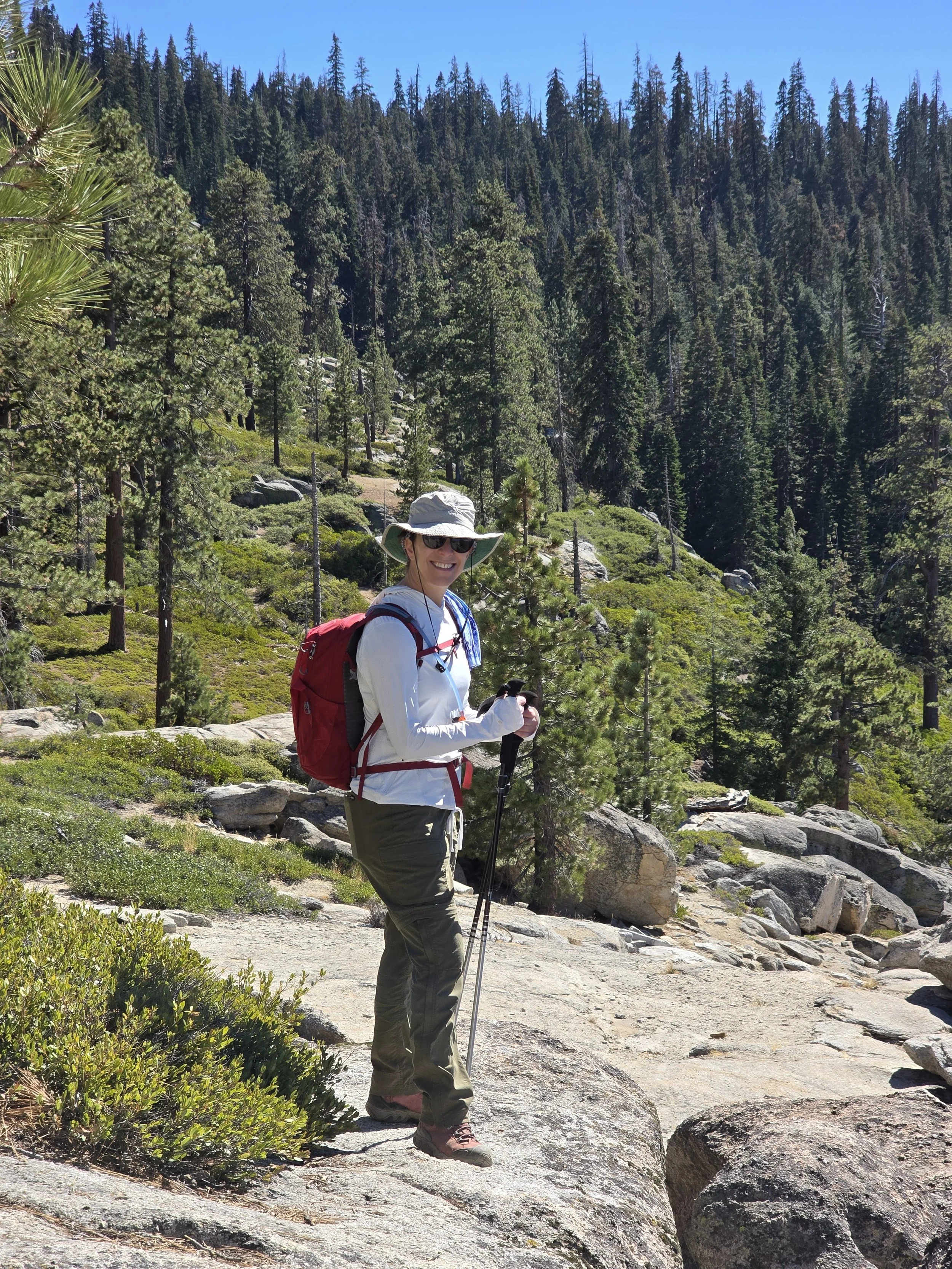 A woman with a white hat, sunglasses, and a red backpack hiking on a rocky trail through a wooded forest with tall pine trees under a clear blue sky.
