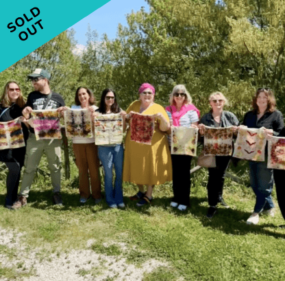 Group of women outdoors holding colorful woven or textile pieces, with trees and blue sky in the background, and a sold-out sign in the corner.