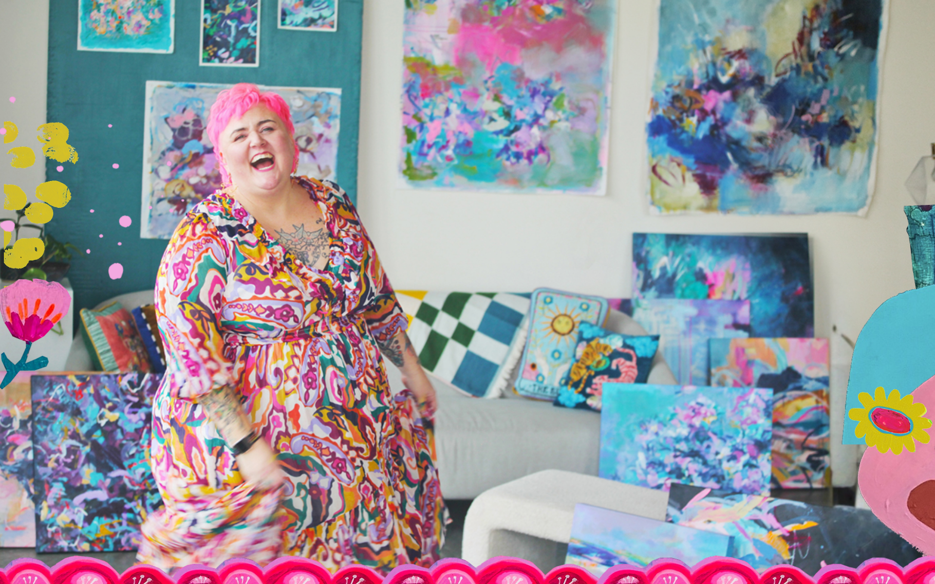 A woman with short pink hair and colorful dress smiling and laughing in an art studio with abstract paintings and colorful cushions on a couch.