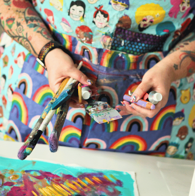 Person holding paintbrushes and tubes of paint, wearing a colorful apron with rainbow and cartoon character designs, in an art studio with a colorful abstract painting on the table.