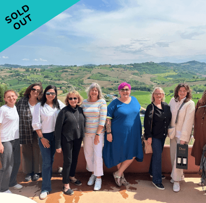 Group of seven women standing outdoors on a balcony with green rolling hills in the background, some wearing sunglasses, dressed casually, one holding a bag, and a