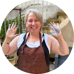 A woman smiling outdoors, showing her hands covered in colorful paint.