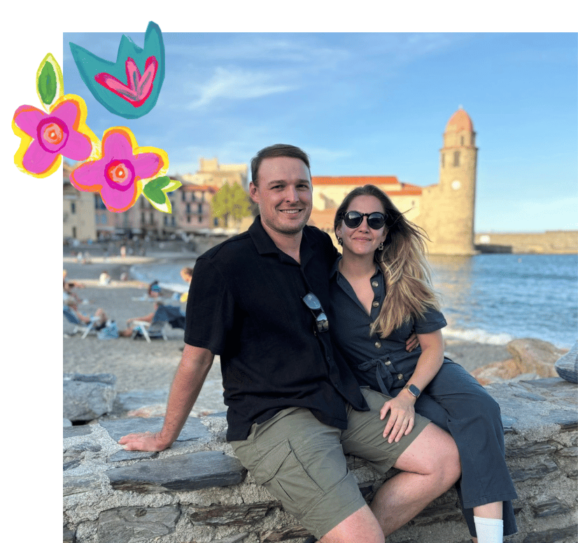 A young man and woman sitting on a stone wall by the water, smiling at the camera on a sunny day. The man is wearing a black shirt and khaki shorts, and the woman is wearing dark sunglasses and a dark shirt with rolled-up sleeves. In the background, there is a historic stone tower and colorful buildings along the waterfront, with a clear blue sky and some people relaxing on the beach.