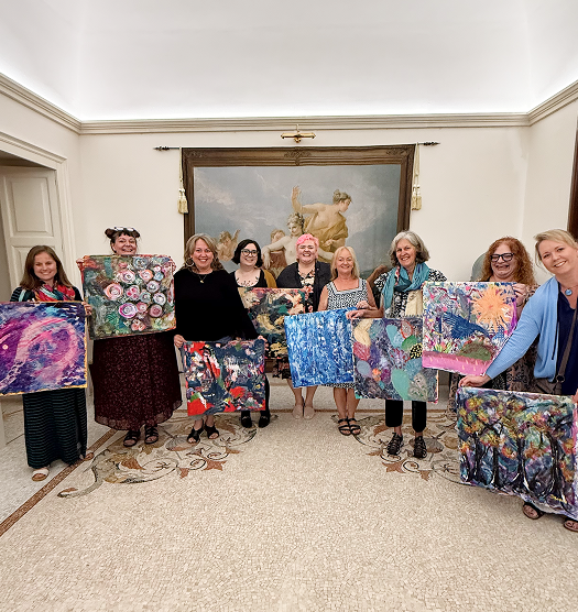 Group of women standing indoors, holding colorful abstract paintings, with a large framed painting on the wall behind them.