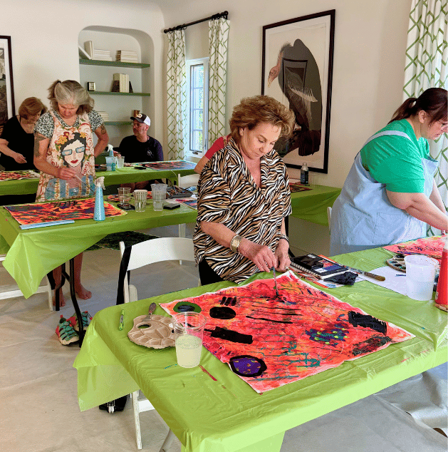 A group of people participating in an art class, creating watercolor paintings on large sheets of paper, with tables covered in green tablecloths and art supplies around.