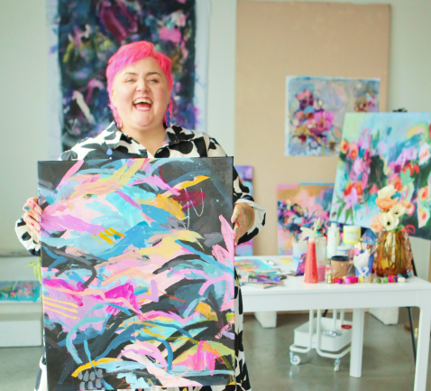 Person with pink hair holding an abstract painting in an art studio.