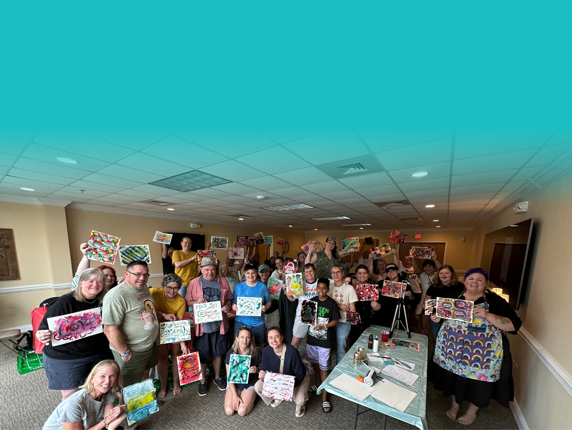 A large group of people in a room holding colorful paintings they created, smiling at the camera during an art event.