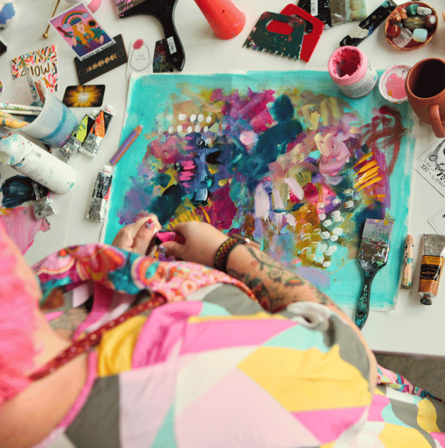 Person creating abstract art on a canvas with colorful paints, surrounded by art supplies and tools on a white table.