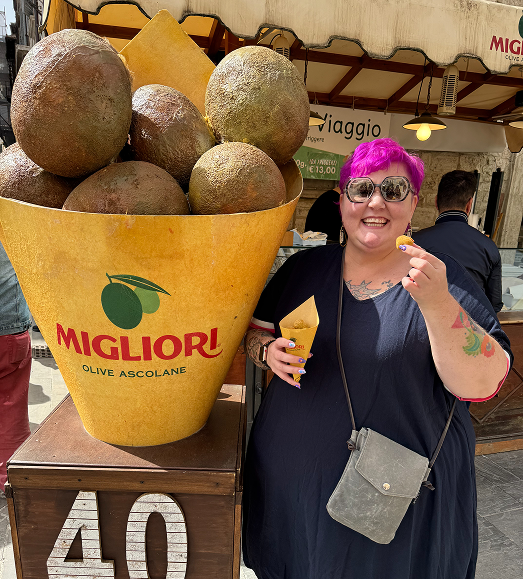 A woman with pink hair, sunglasses, and a navy dress smiling and holding an olive and a cone, standing next to a giant olive cone display with the logo 'Migliori Olive Ascolane' at an outdoor market.