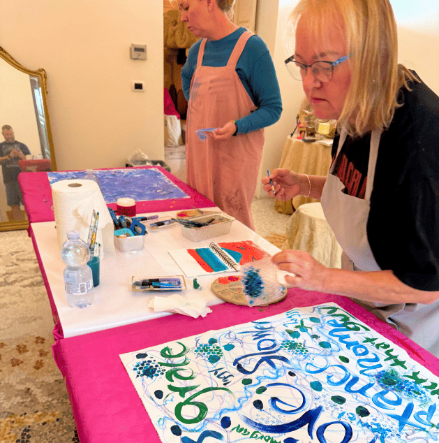 Two women decorating banners at a table with art supplies, one woman is painting while the other looks on, in a room with a mirror and a beige carpet.