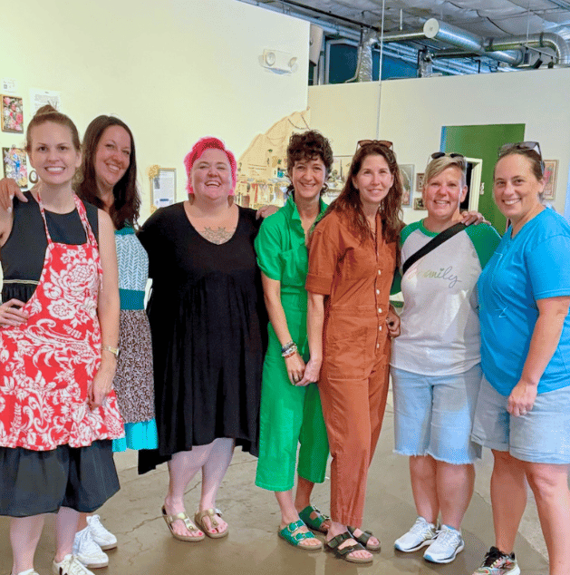 Seven women standing together in a room, smiling for a group photo. The background has light-colored walls and artwork.